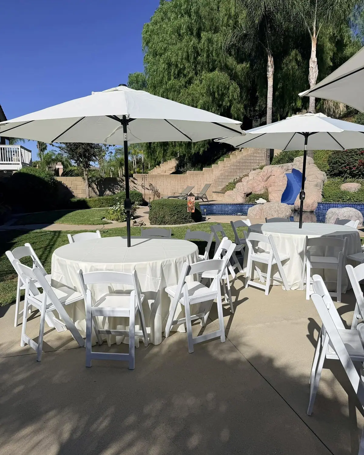 Round tables with ivory tablecloths, white resin folding chairs and canvas patio umbrellas set up at a backyard event