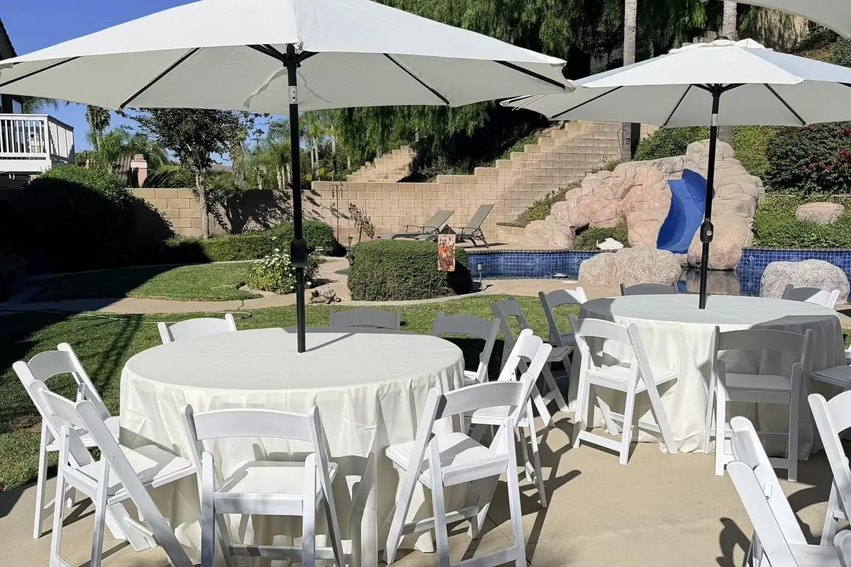 Round tables with ivory tablecloths, white resin folding chairs and canvas patio umbrellas set up at a backyard event