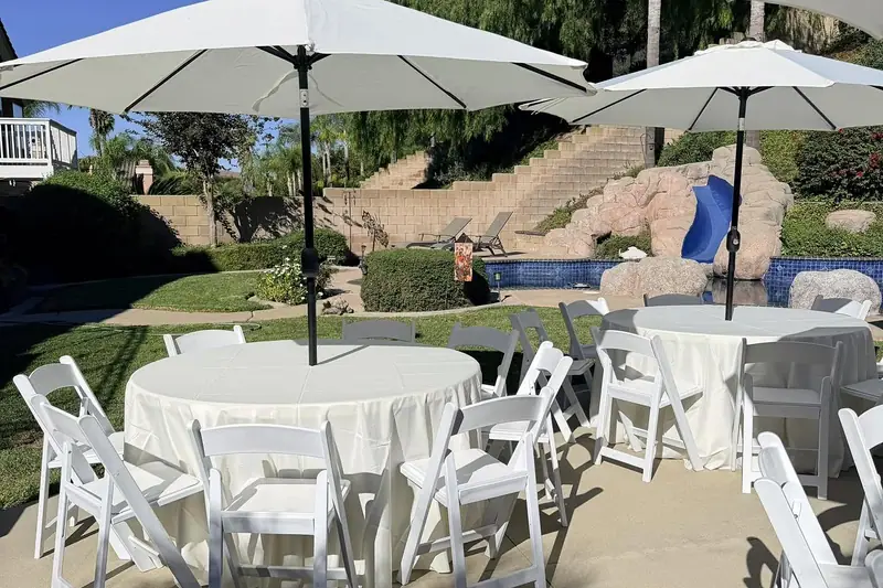 Round tables with ivory tablecloths, white resin folding chairs and canvas patio umbrellas set up at a backyard event
